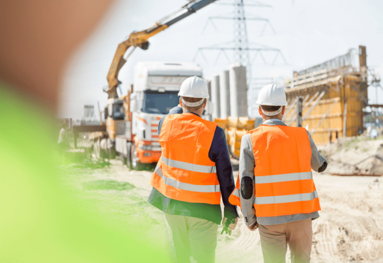 two men in neon orange vests facing away from the camera and pointing to a construction site.