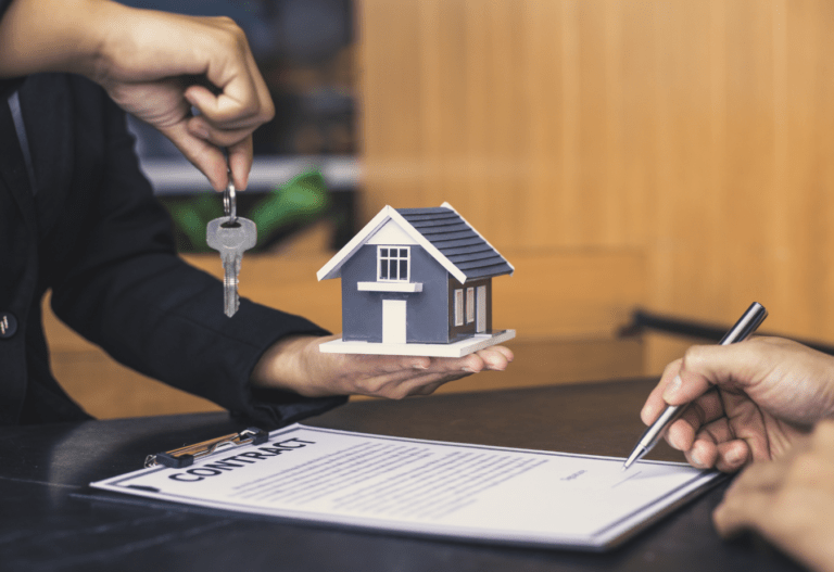 a person on the left handing keys to the person on the right who is signing a contract, a miniature house sits in the background